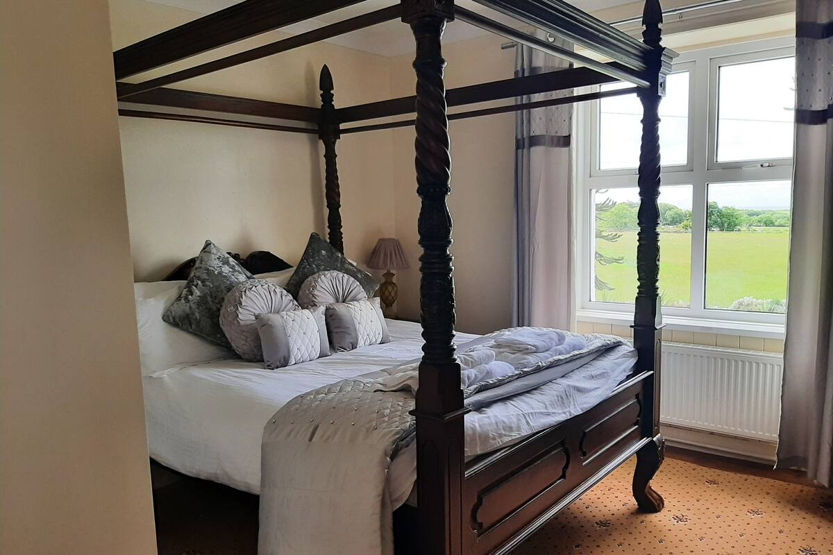 Un lit à baldaquin avec un couvre-lit blanc et de nombreux oreillers, un plancher en bois et une fenêtre donnant sur un paysage verdoyant.
