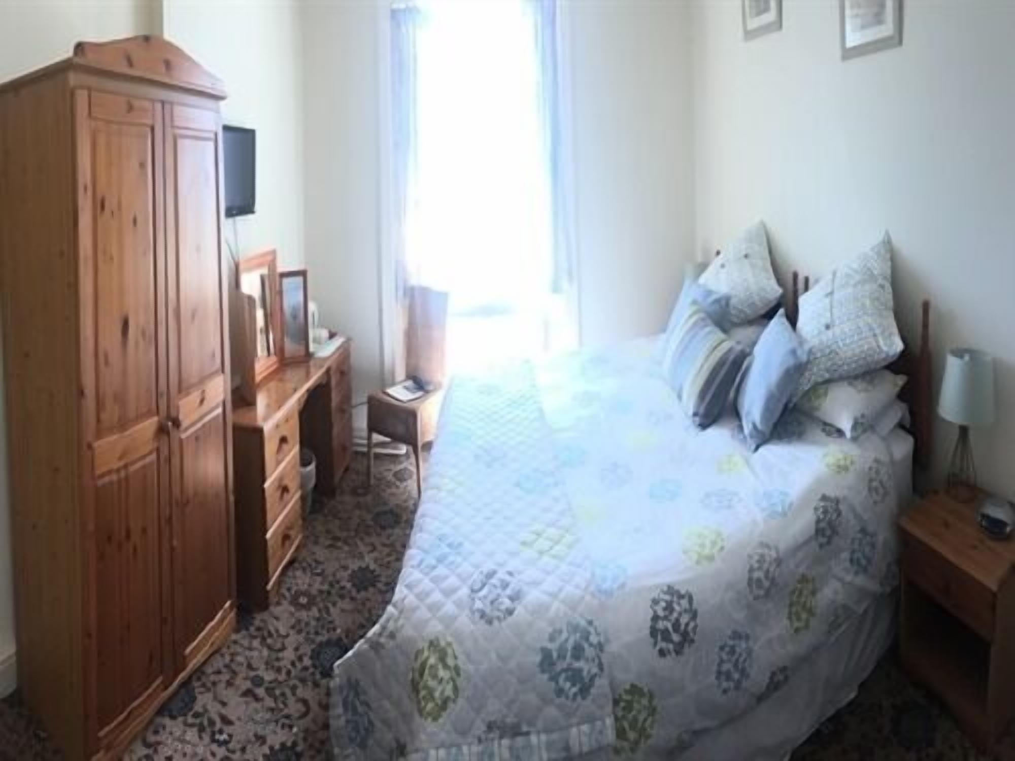 Un dormitorio con una cama, un armario de madera, un escritorio con silla, una mesita de noche con lámpara y una ventana con cortinas.
