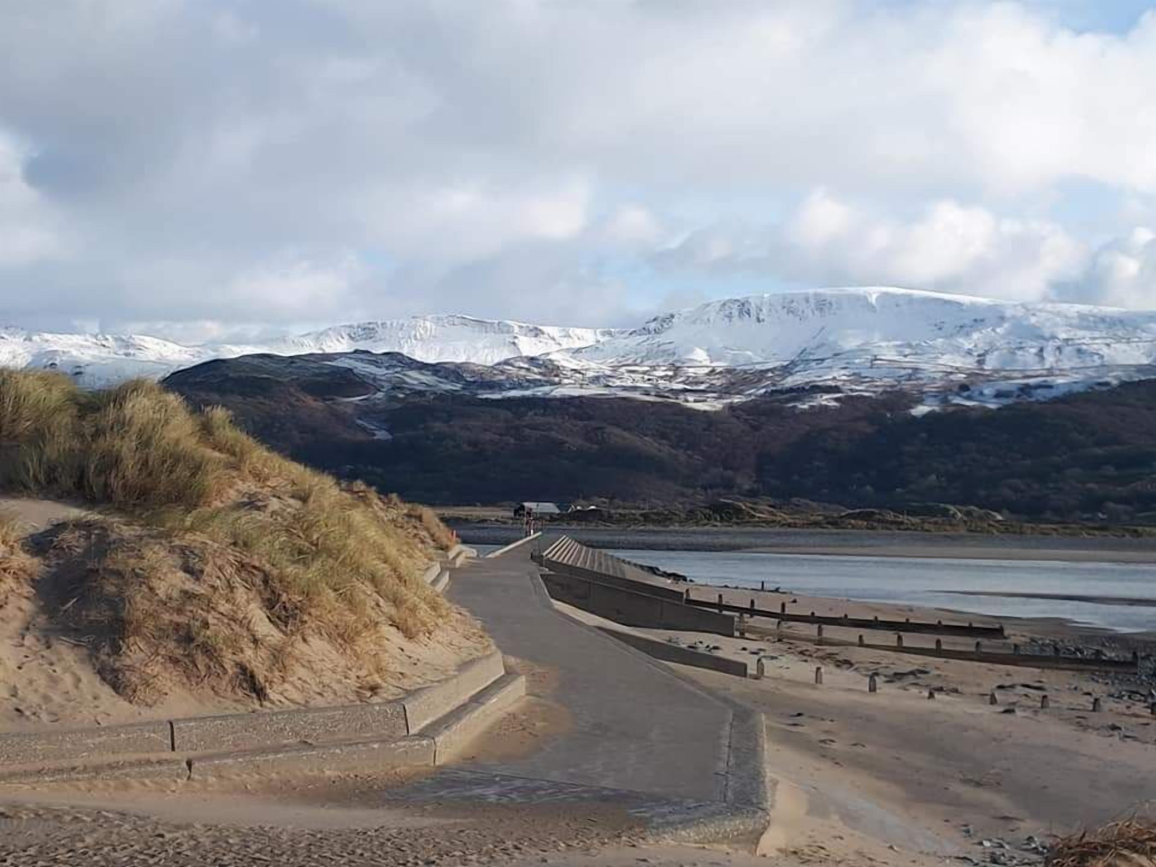 Une route côtière bordée d’une barrière de sécurité, qui mène à une plage d’où l’on peut admirer des montagnes enneigées en arrière-plan.