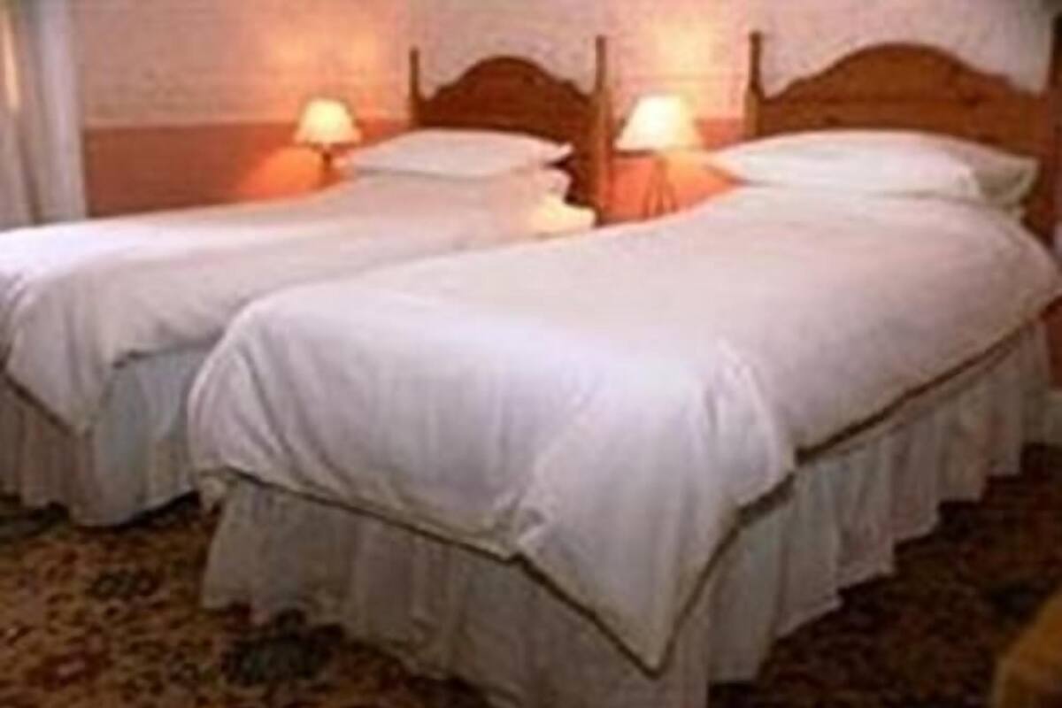 Une chambre d’hôtel avec deux lits simples, chacun doté d’une tête de lit en bois et d’une lampe de chevet. La chambre est dotée d’un sol recouvert de moquette et d’un mur uni.