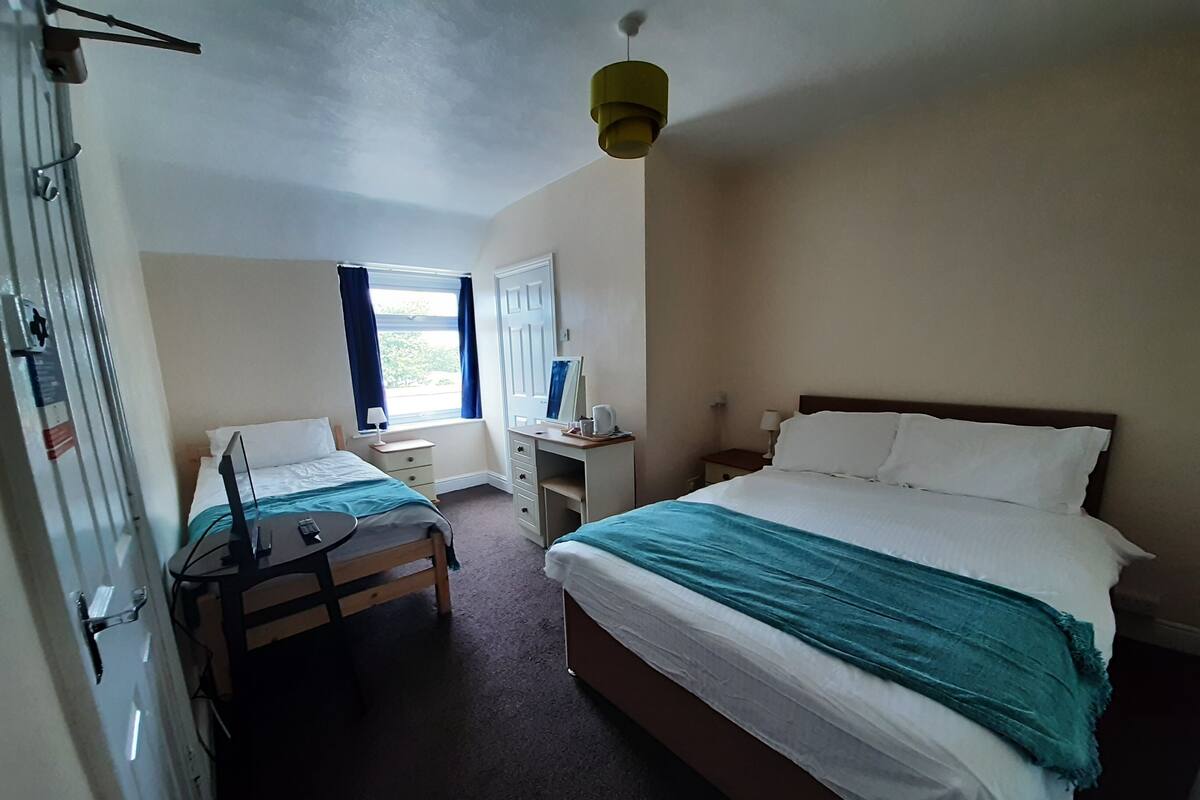 Une chambre d’hôtel comprenant un lit, un bureau, une chaise, une lampe de bureau, une fenêtre avec des rideaux et un luminaire au plafond.