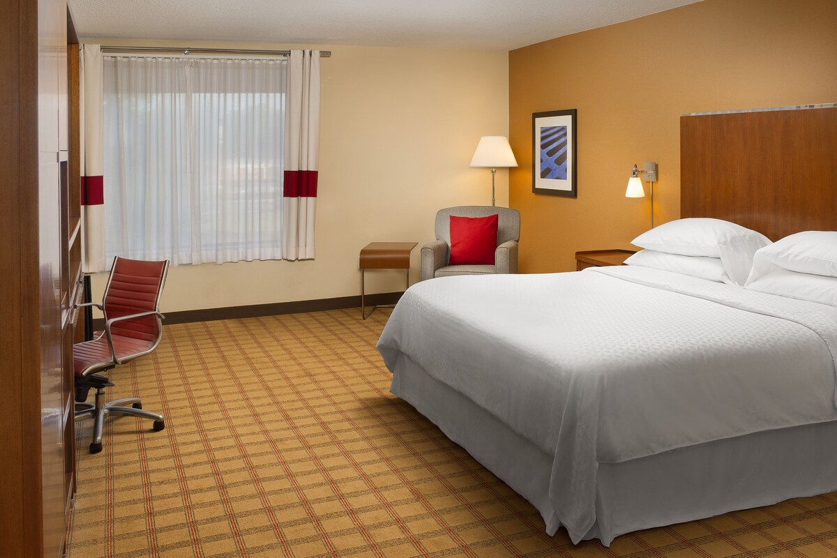 Habitación de hotel con cama, una silla, escritorio, lámpara y ventana con cortinas.