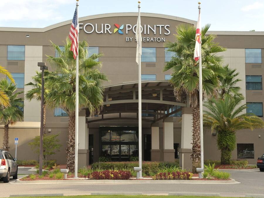 A hotel entrance with a sign reading "Four Points by Sheraton," palm trees, and a parking area.