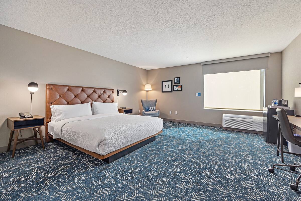 Una habitación de hotel con una cama grande, un escritorio, una silla y un ventanal amplio.