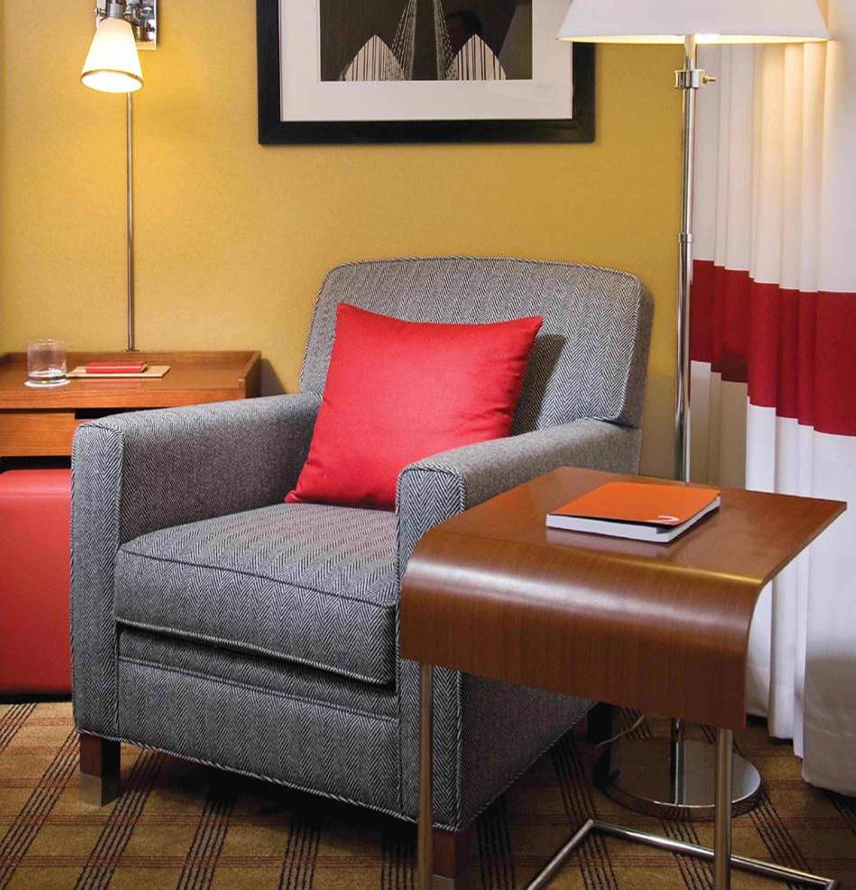 Una habitación de hotel con un sillón gris y un cojín rojo, una mesita de noche de madera con un libro, una lámpara de pie y un cuadro en la pared.