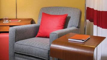 Una habitación de hotel con un sillón gris y un cojín rojo, una mesita de noche de madera con un libro, una lámpara de pie y un cuadro en la pared.