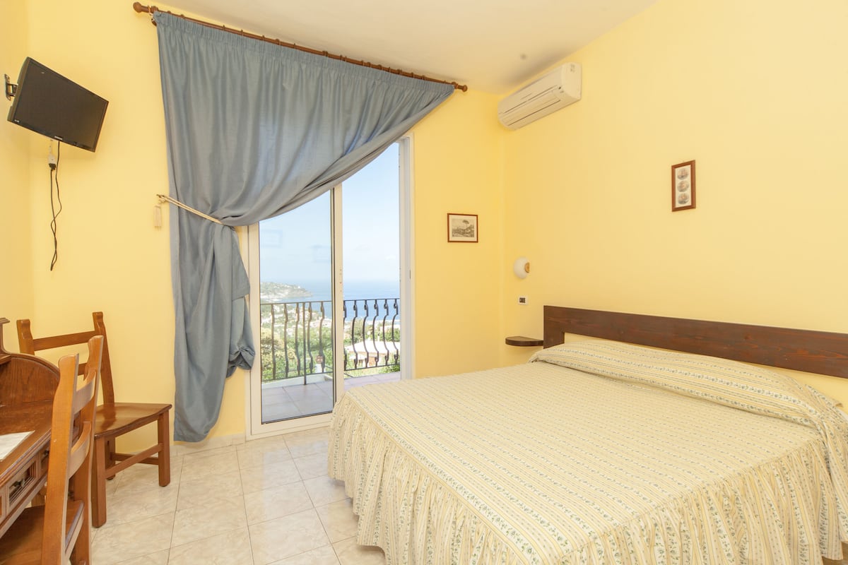 Een slaapkamer met een bed, een bureau, een stoel, een televisie en een balkon met uitzicht.