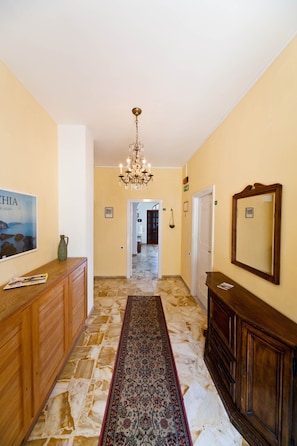 A hallway with a chandelier, a wooden dresser, a mirror, and a patterned rug. - Hotel Ape Regina (Casamicciola Terme)