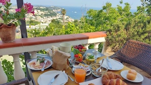 Mesa posta com itens de café da manhã, incluindo croissants, doces e uma saladeira de frutas, com vista para a paisagem costeira.