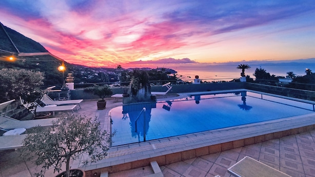 Piscina com vista para o pôr do sol, cadeiras de descanso e plantas em vasos.