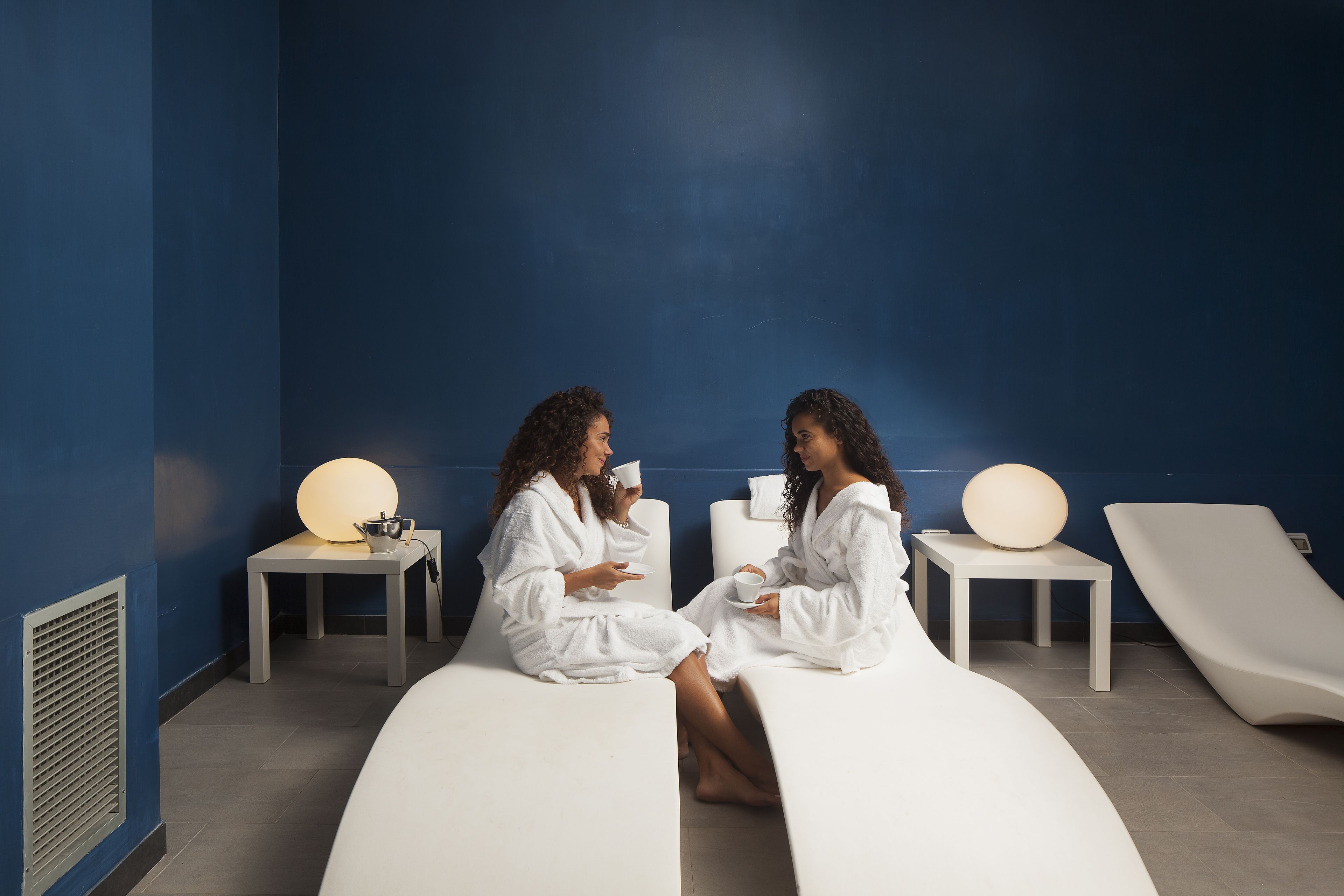Dos personas vestidas de blanco sentadas en un banco curvo en un spa con paredes azules, una mesa blanca y una silla blanca.
