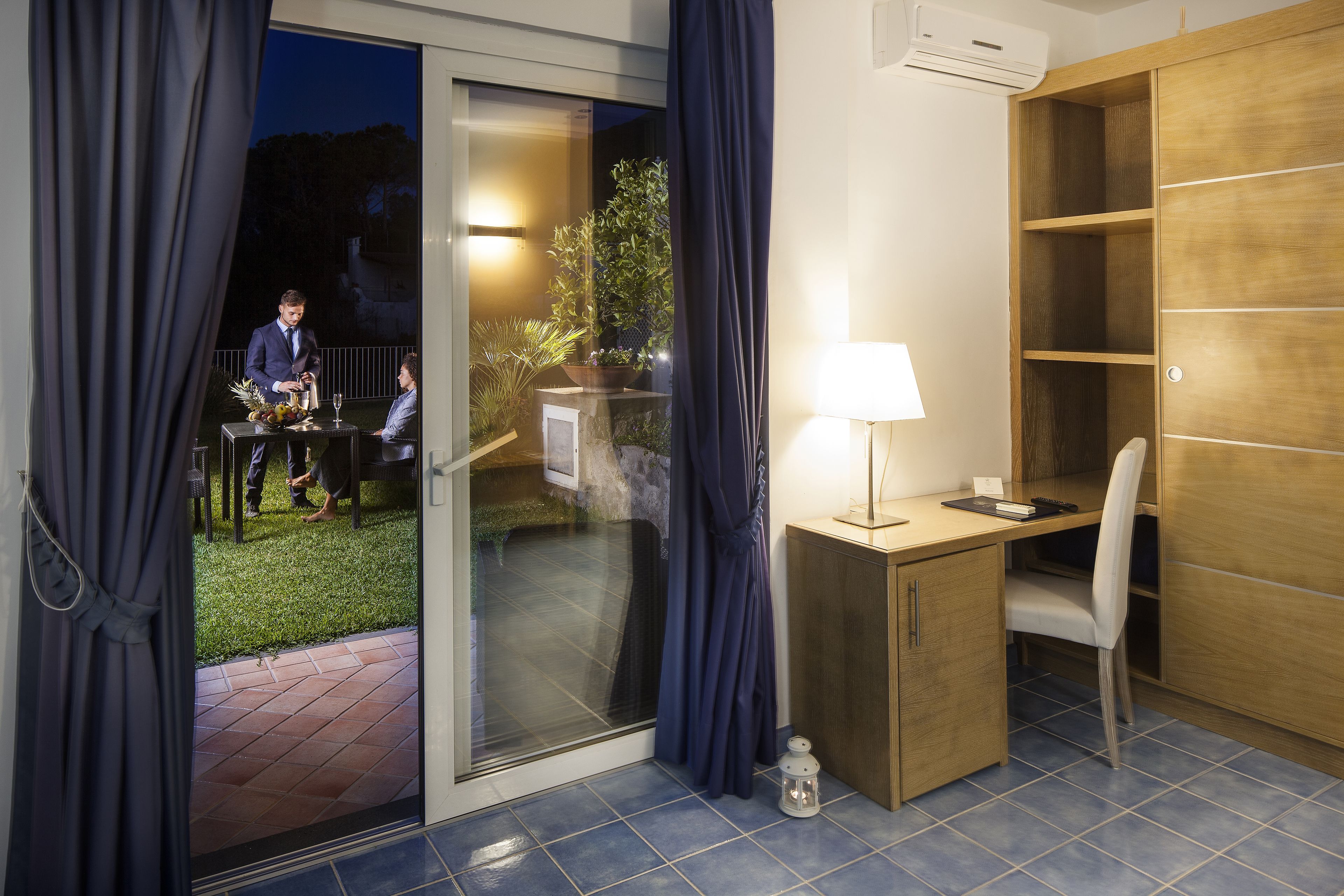 A room with a sliding glass door leading to a balcony, a desk with a chair, a small table with a lamp, and a wardrobe.