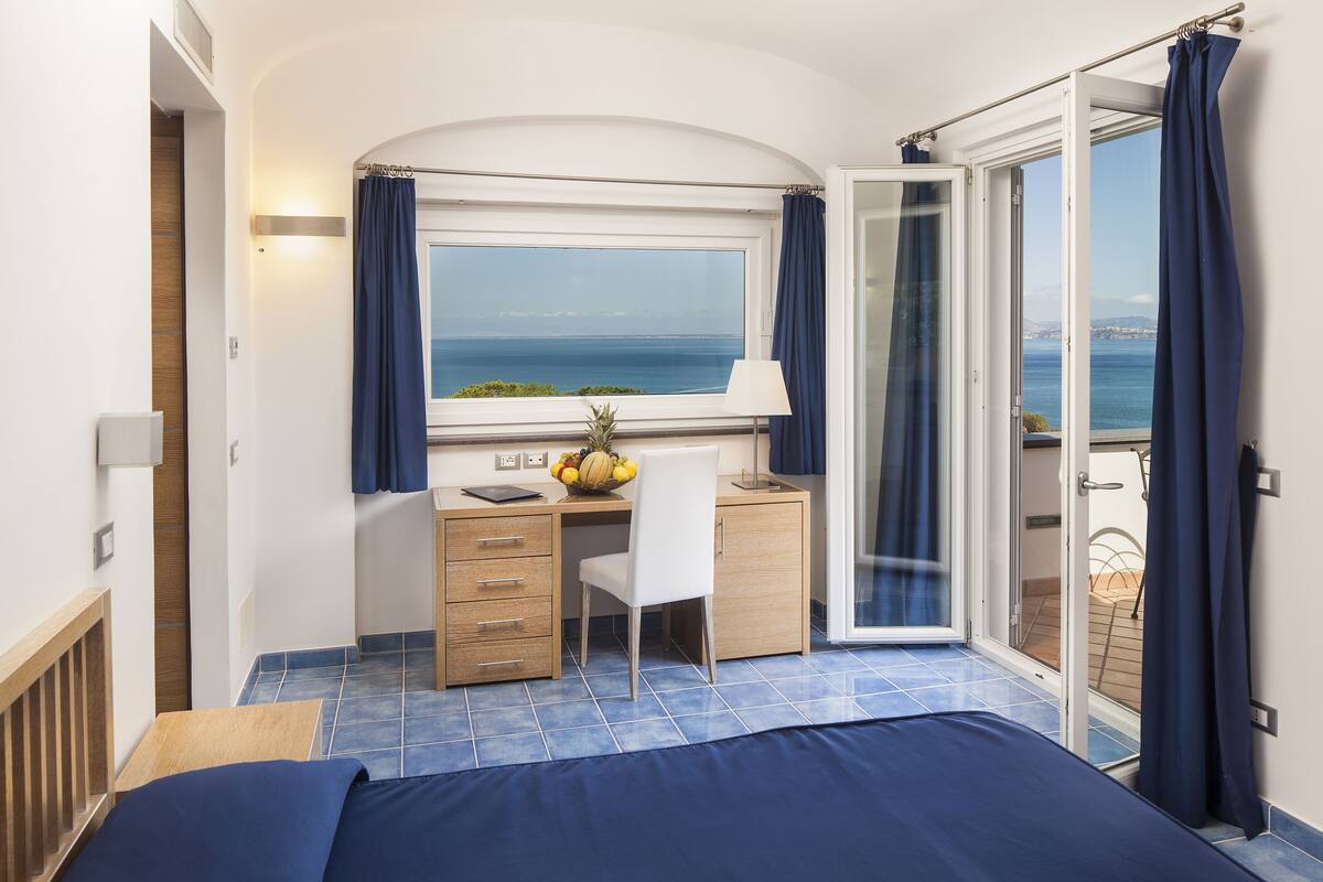 A bedroom with a bed, a desk, a chair, a window with curtains, and a view of the sea.