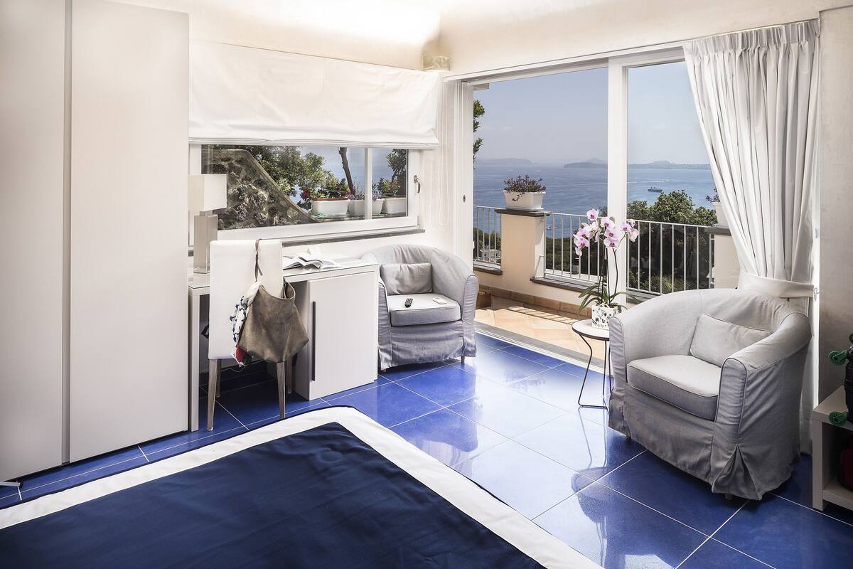 A balcony with a view of the sea, a white sofa, a small table, and a potted plant.