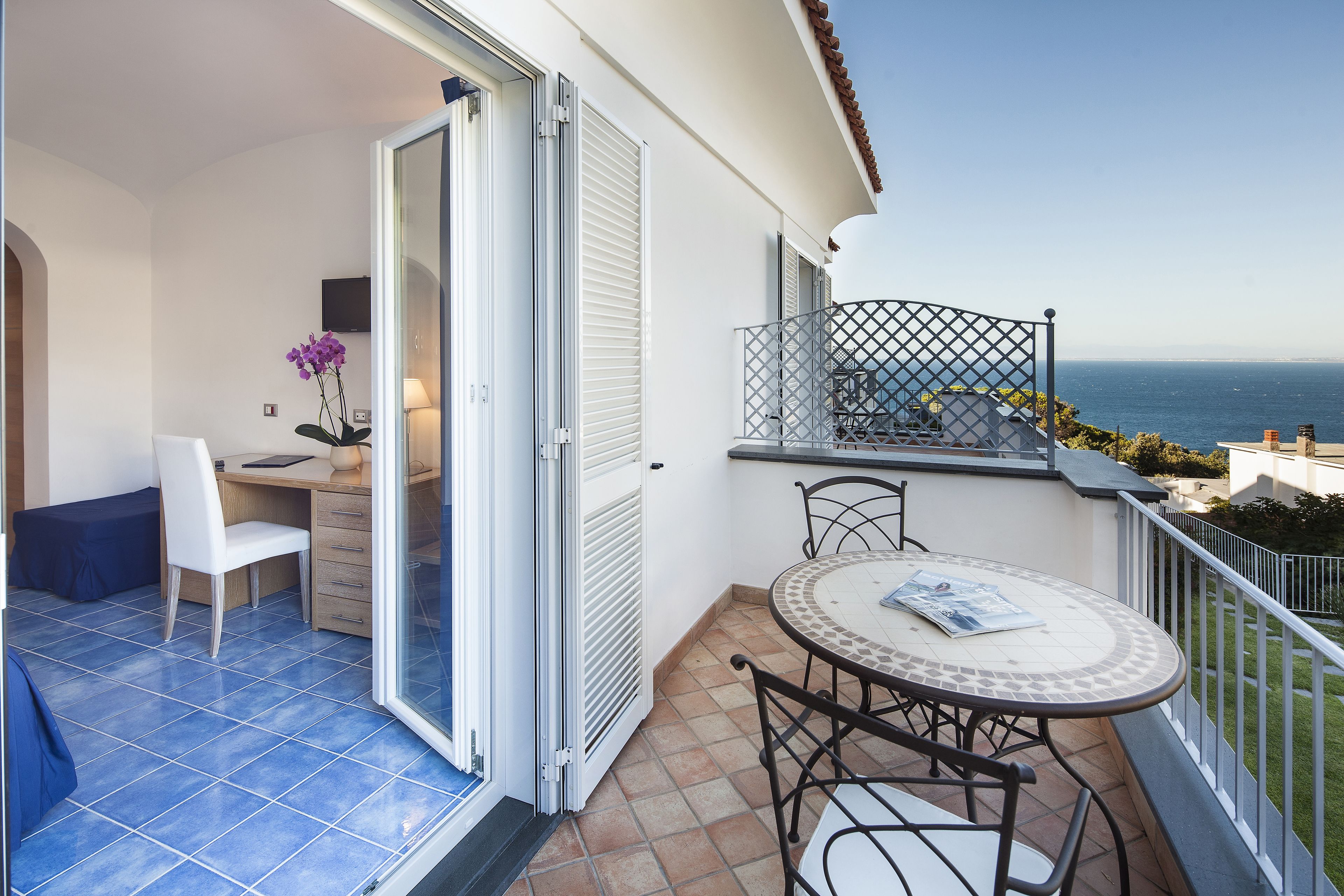 A balcony with a table and chairs, a chair, a desk with a lamp, and a view of the sea.