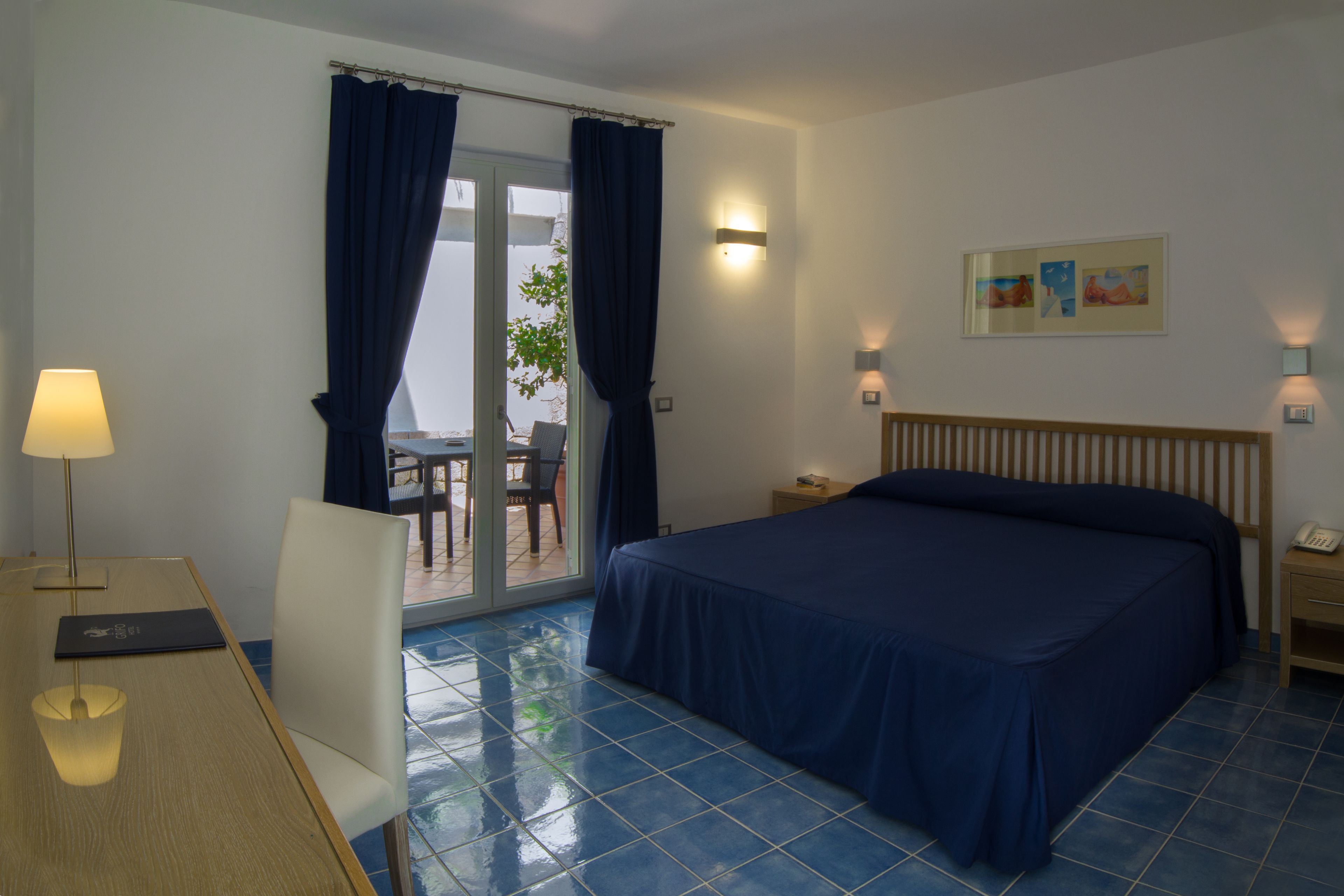 Une chambre d’hôtel avec un lit, un bureau doté d’une lampe, une chaise, une table de chevet et un tableau au mur.
