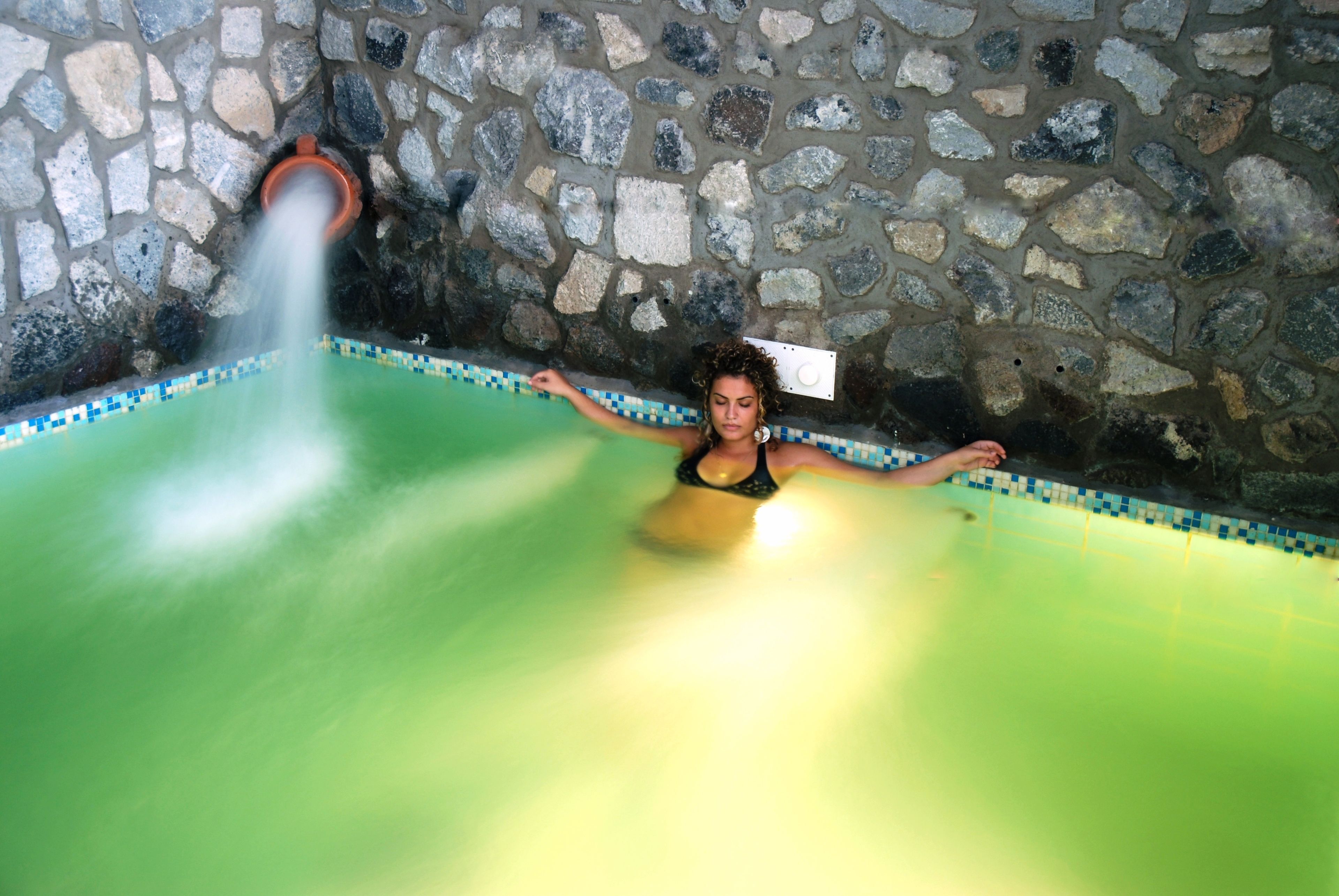 A person in a stone-walled room with a green pool, a water spout, and a tiled border.