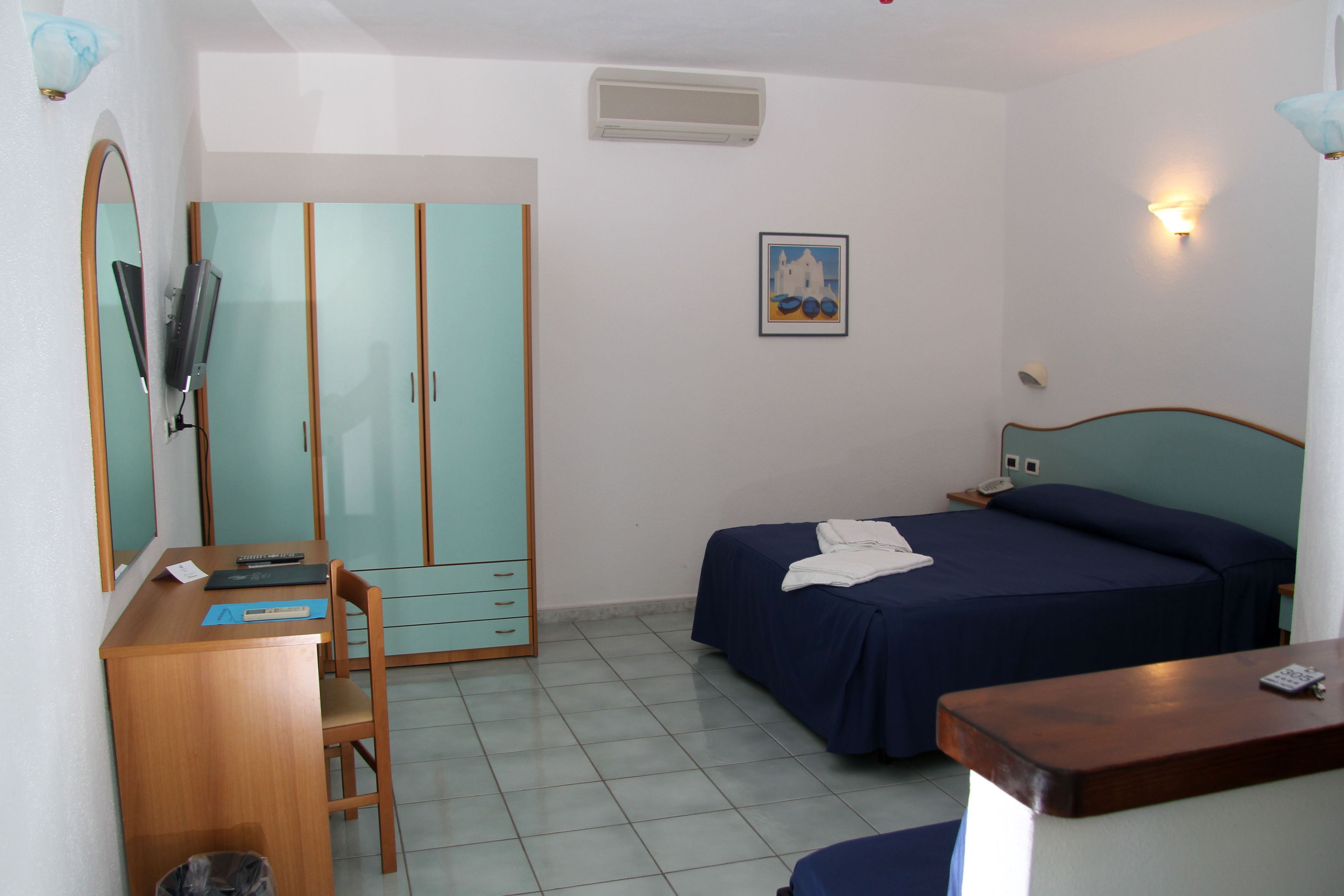 Habitación de hotel con cama, escritorio con silla, armario, espejo y televisor montado en la pared.