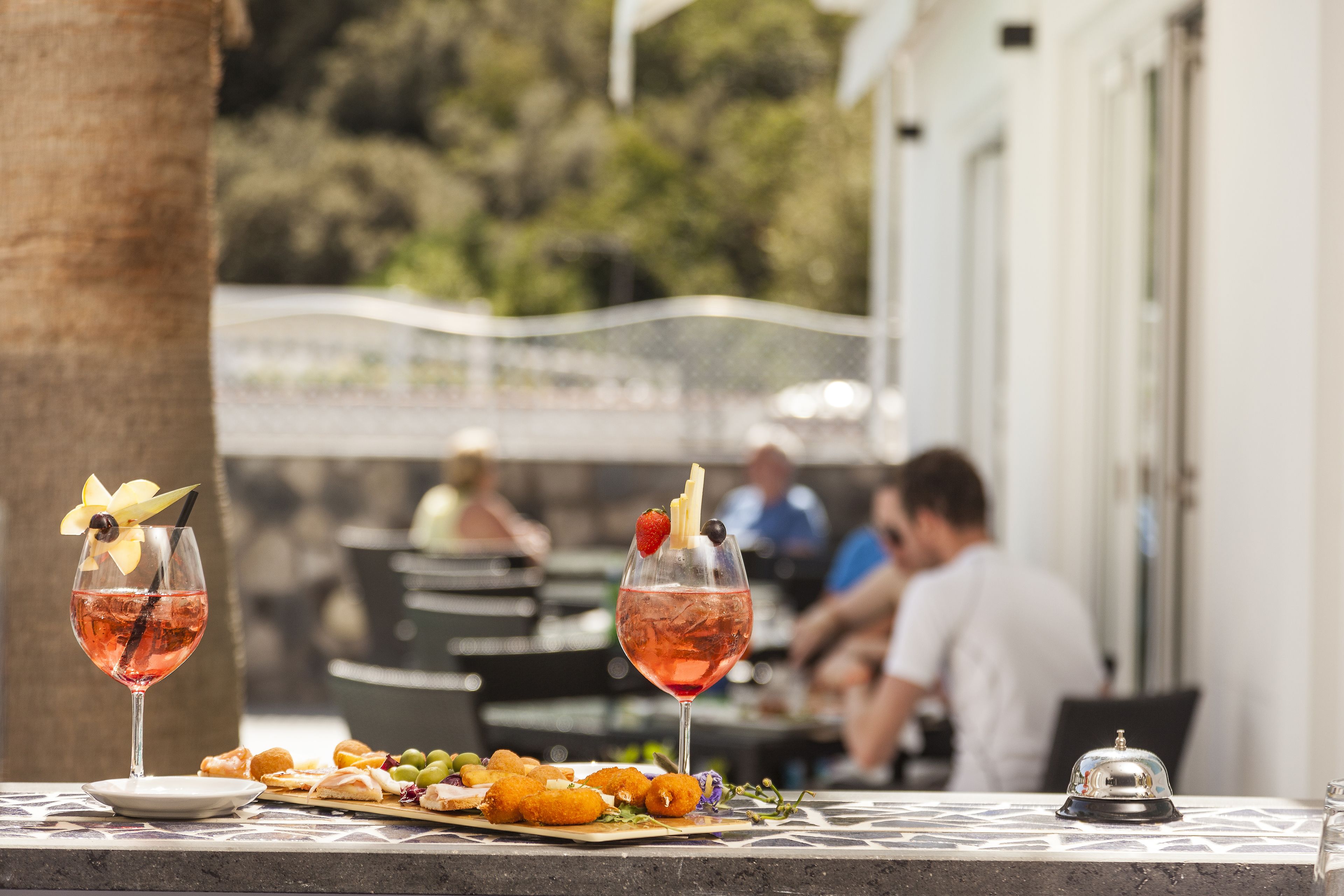 Une table sur laquelle se trouvent deux verres contenant une boisson rose, une assiette de hors-d'œuvre et une clochette, le tout posé sur une table dont le plateau est décoré de motifs.