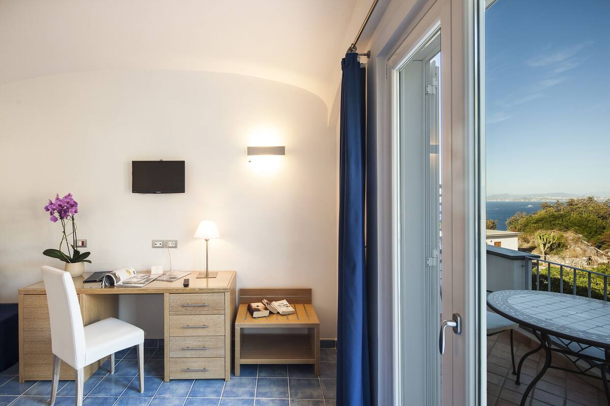 A bedroom with a desk, chair, bedside table, and a view of the outdoors.