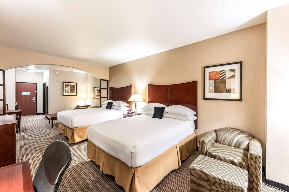 A hotel room with two beds, a desk, a chair, and a lamp.