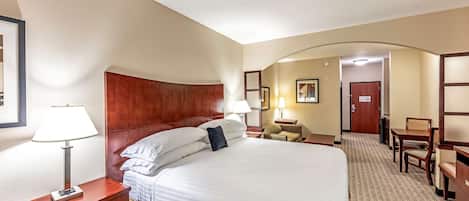A hotel room with a large bed, a wooden headboard, a bedside table with a lamp, a desk with a chair, and a door leading to another room.