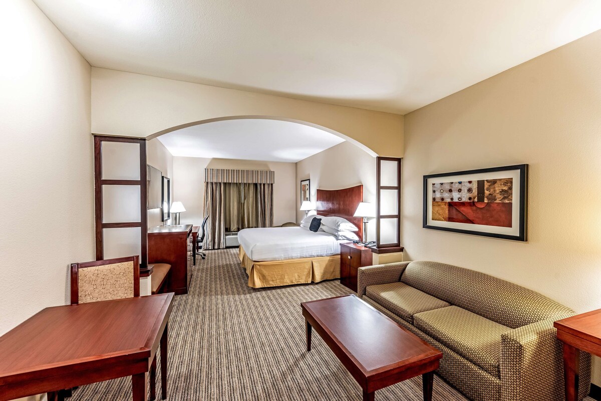 A hotel room with a bed, a sofa, a desk, and a chair. There is a painting on the wall and a window with curtains.