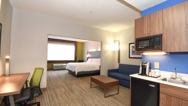 A hotel room with a bed, a desk, a chair, a sofa, a kitchenette with a microwave, and a window.