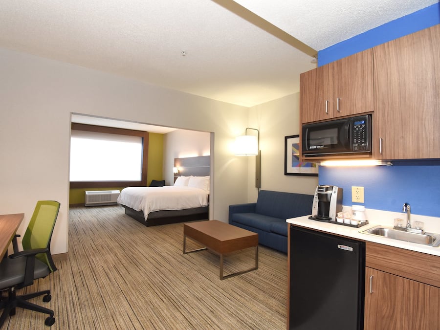 A hotel room with a bed, a desk, a chair, a sofa, a kitchenette with a microwave, and a window.