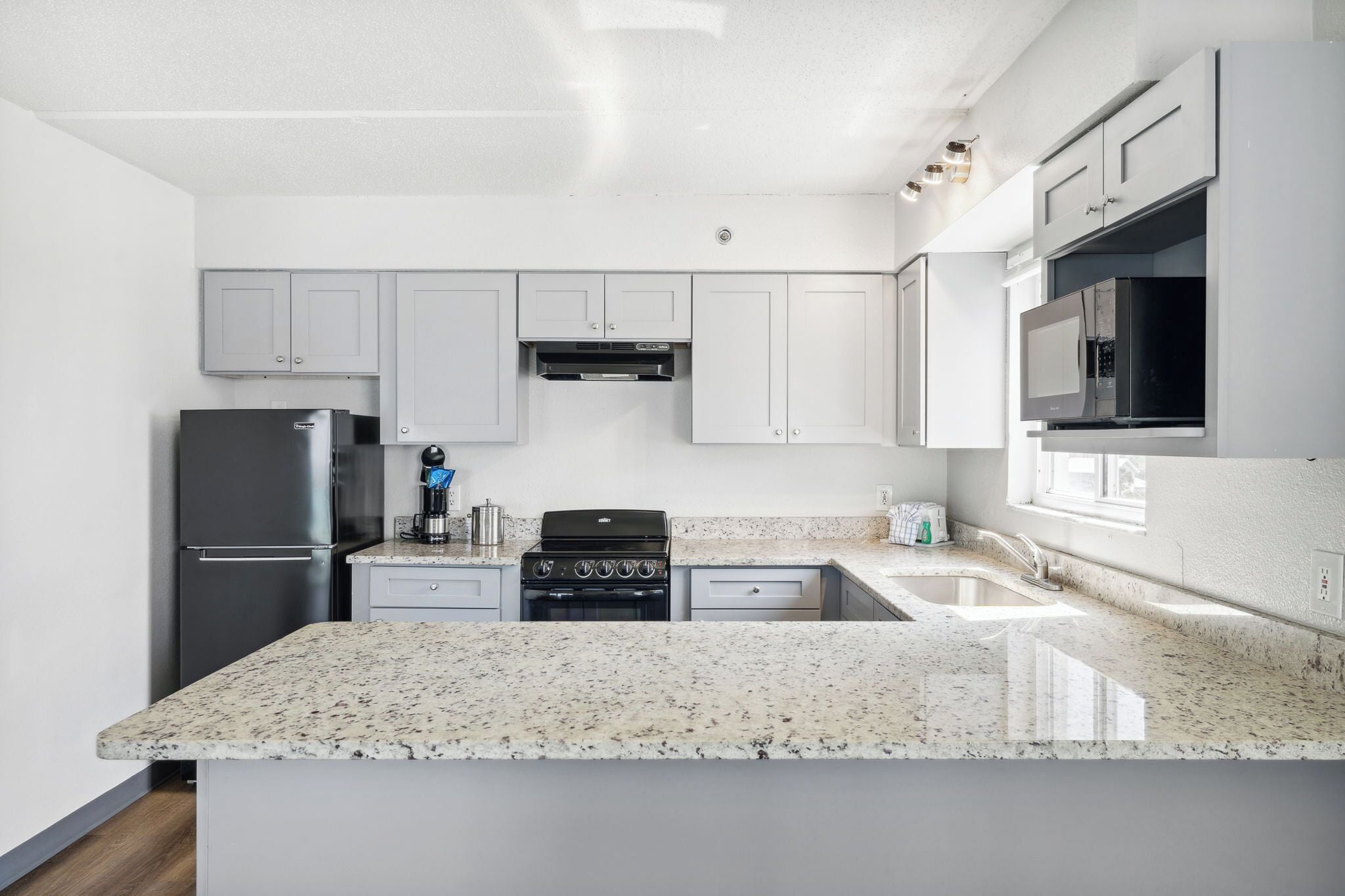 A modern kitchen with white cabinetry, a granite countertop, a stainless steel refrigerator, a stove, and a microwave.