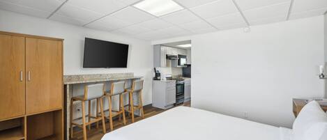 A hotel room with a bed, a desk with chairs, a TV, a kitchenette, and a wooden wardrobe.