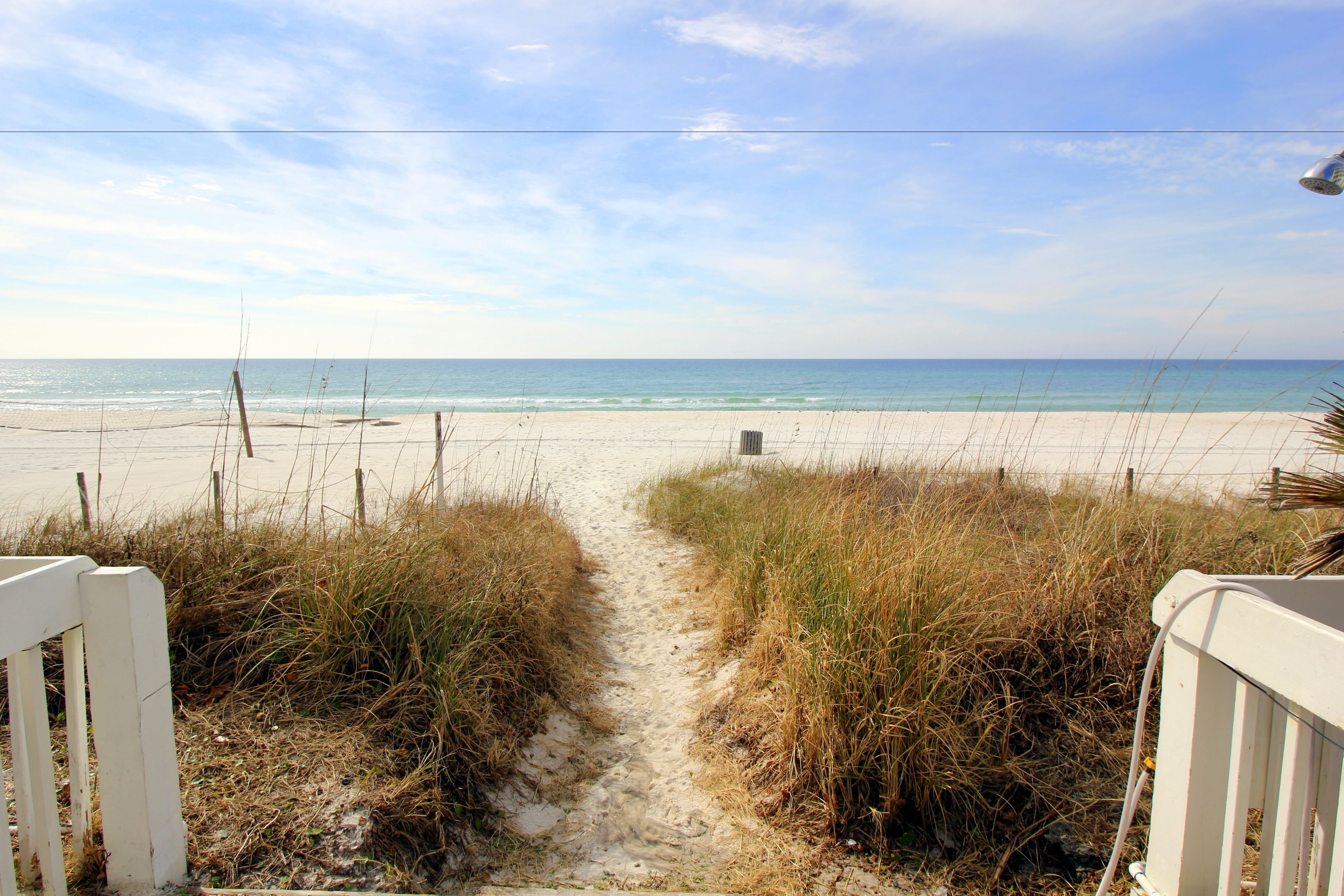 A sandy beach path leading to the ocean, with a white fence on one side and tall grass on the other.