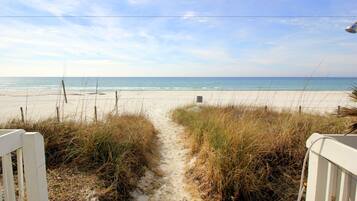A sandy beach path leading to the ocean, with a white fence on one side and tall grass on the other.