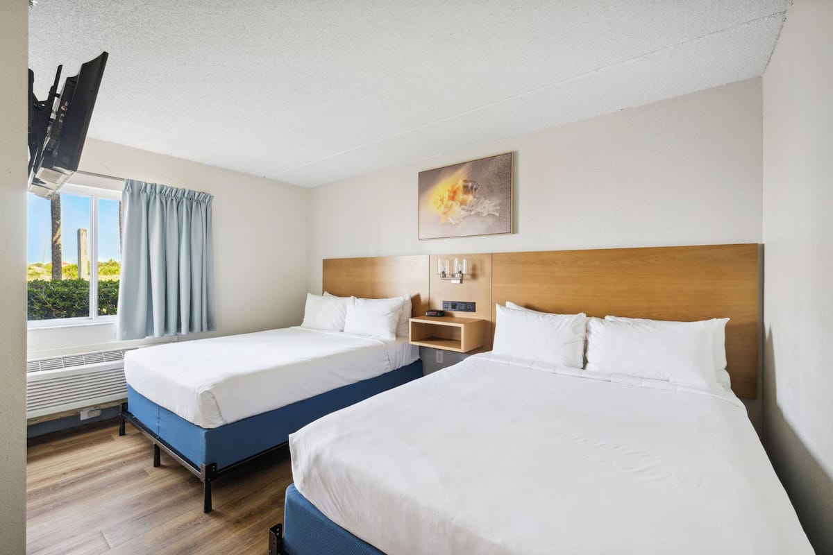 Habitación de hotel con dos camas, cabecera de madera, mesita de noche con reloj y ventana con cortinas.