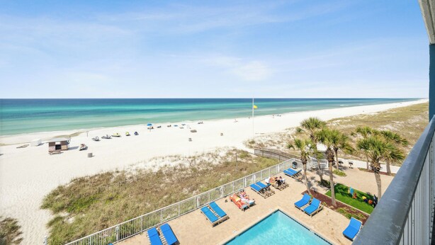 A beach view from a balcony with a pool, lounge chairs, and a lifeguard tower.
