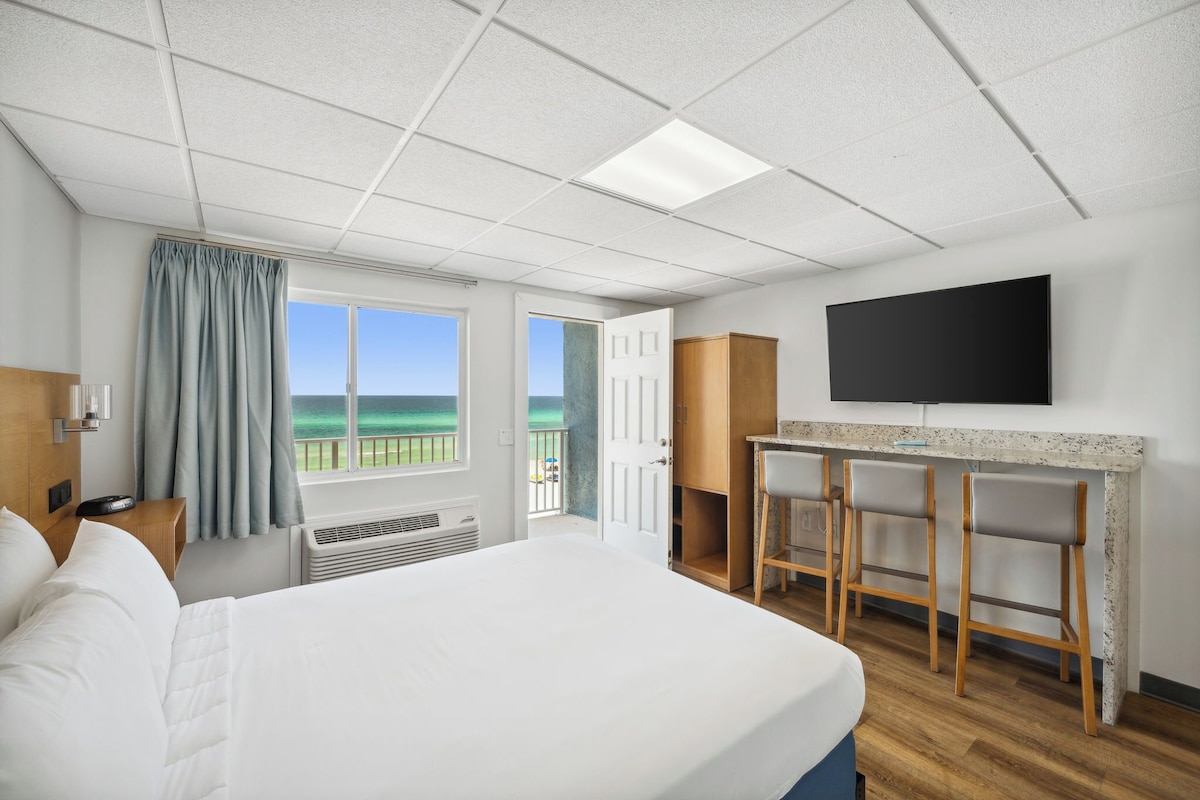 Hotelkamer met een bed, een bureau met barstoelen, een televisie, een raam met uitzicht op zee en een deur naar een balkon.