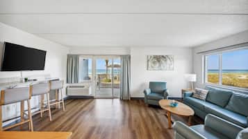 A modern hotel room with a kitchen, dining area, sofa, and a balcony with a view of the beach.