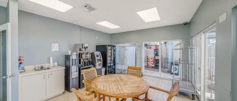 A dining area with a round wooden table and chairs, a refrigerator, a coffee machine, and a vending machine.