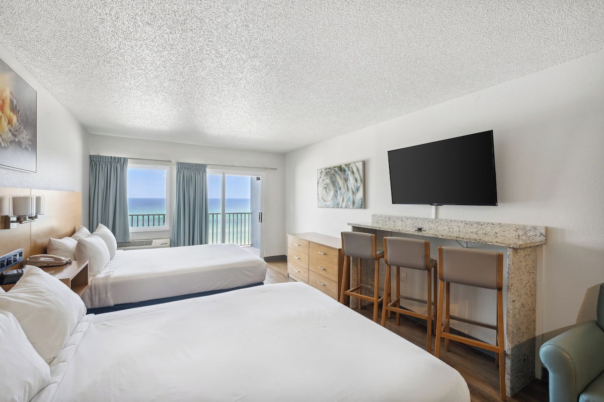Una habitación de hotel con dos camas, un escritorio con sillas, un televisor y vista al mar.