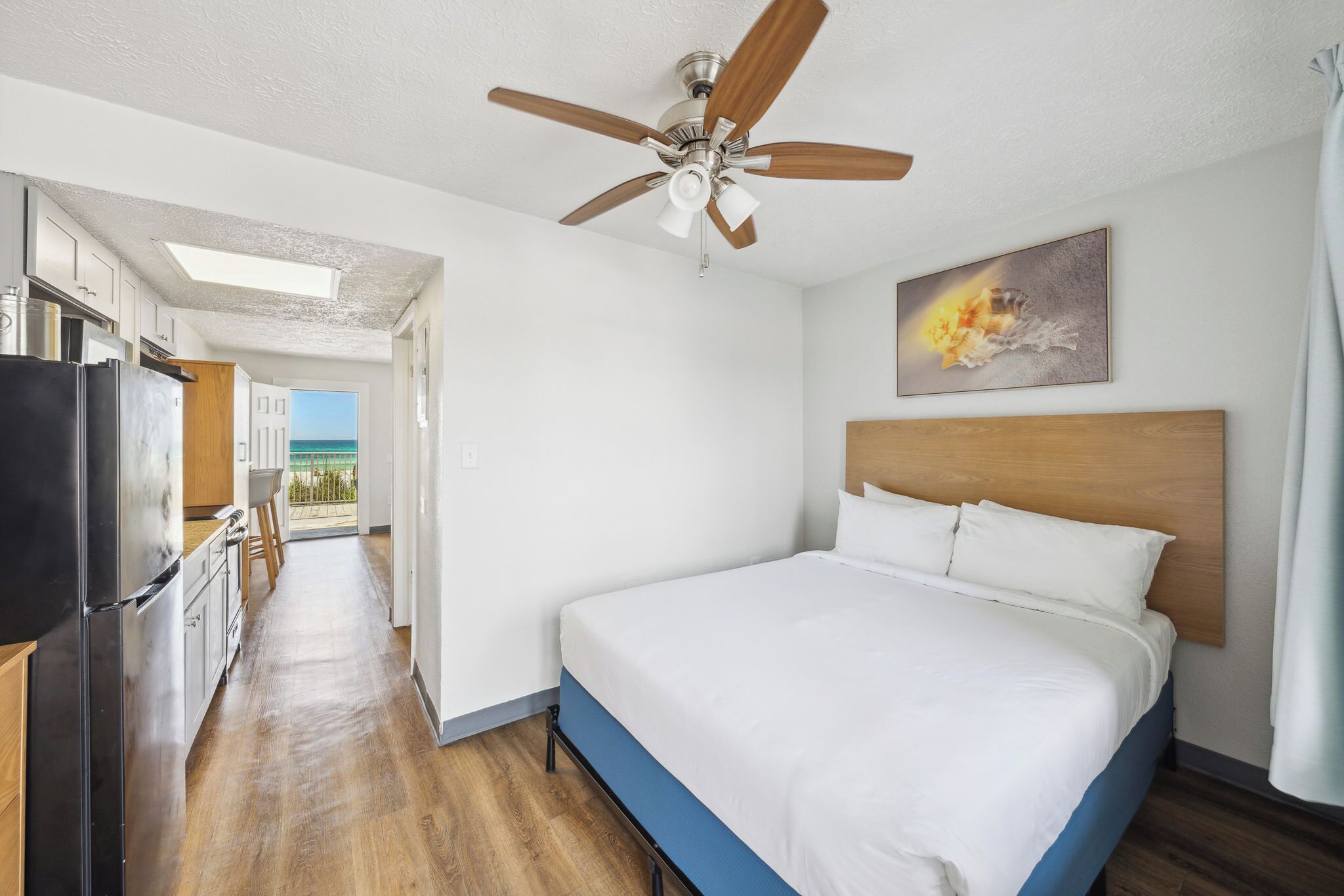 A hotel room with a bed, a ceiling fan, a painting, and a view of the outside.
