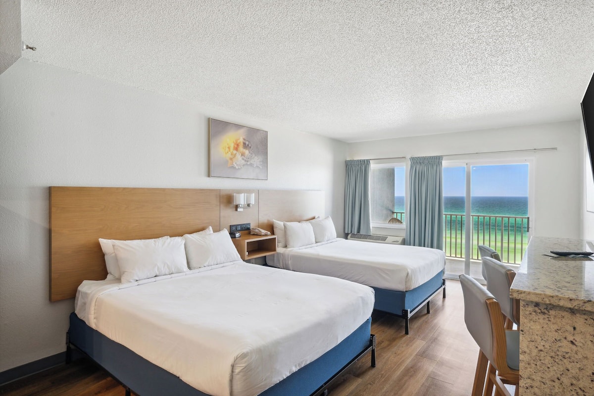 Habitación de hotel con dos camas, un escritorio, una silla, un cuadro y un balcón con vistas.