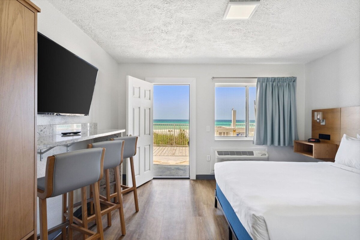 Een hotelkamer met een bed, een bureau met stoelen, een televisie, een raam met uitzicht op het strand en een nachtkastje met een lamp.