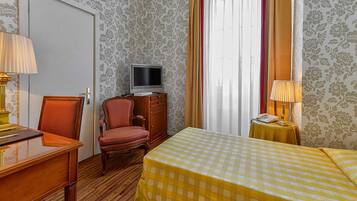 Ein Hotelzimmer mit Holztisch, Fernseher, Bett mit kariertem Überwurf und kleiner Tisch mit Lampe.