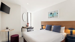 A hotel room with a large bed, a desk with a laptop, a small round table, a TV mounted on the wall, and a painting above the bed. - Quest Ivanhoe (Ivanhoe)