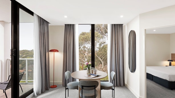 A modern hotel room with a round dining table, four chairs, a large window with curtains, a floor lamp, and a bed with a nightstand. - Quest Ivanhoe (Ivanhoe)
