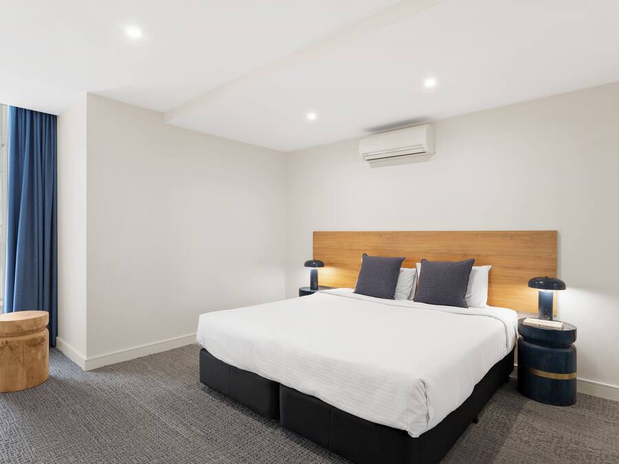 A modern hotel room with a large bed, wooden headboard, two bedside tables with lamps, a small wooden stool, and a large window with blue curtains.