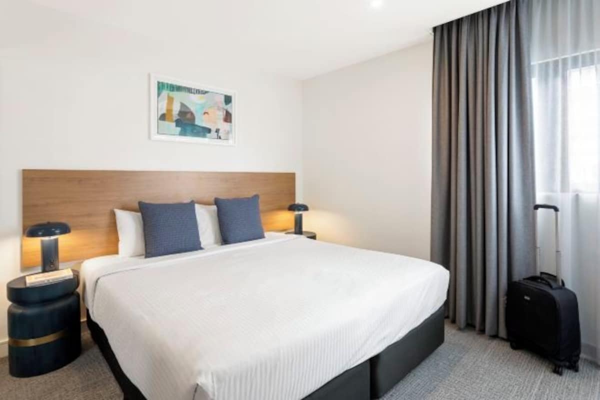 A hotel room with a large bed, two bedside tables with lamps, a headboard, a painting on the wall, and a window with curtains.