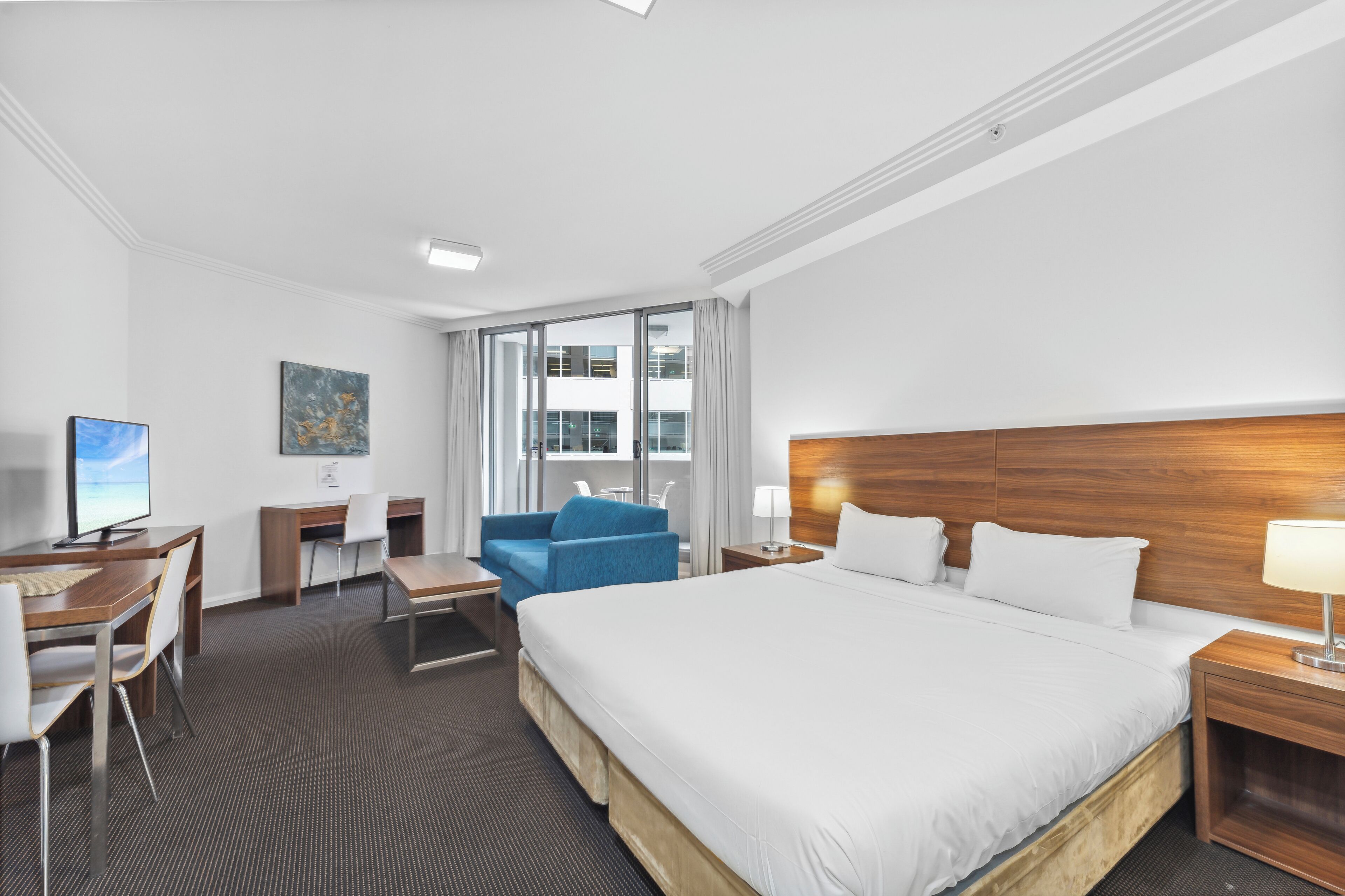 A hotel room with a large bed, a desk with a computer, a blue chair, and a view of the city through large windows.
