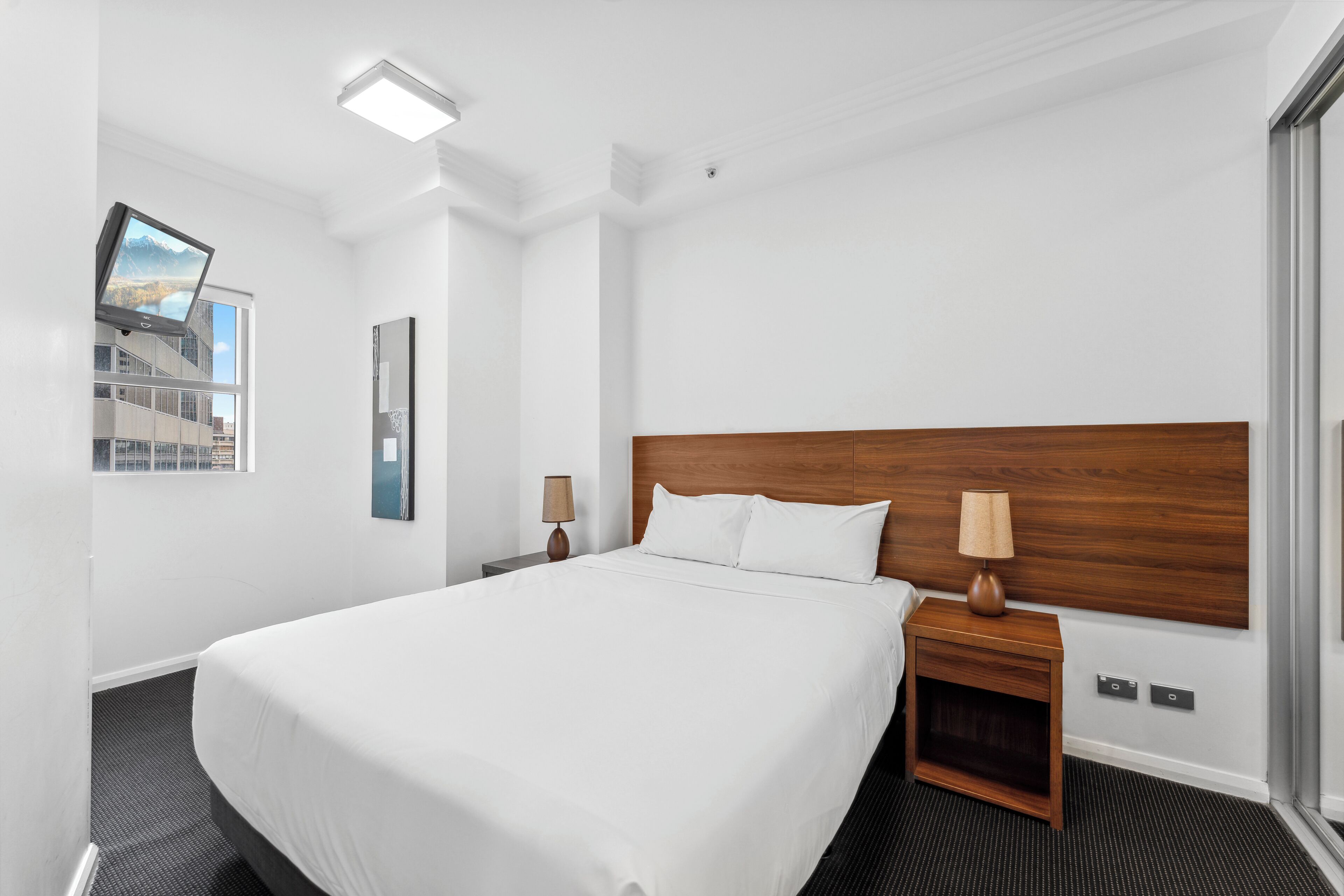 A hotel room with a large bed, wooden headboard, two bedside tables with lamps, a window with a cityscape view, and a framed picture on the wall.