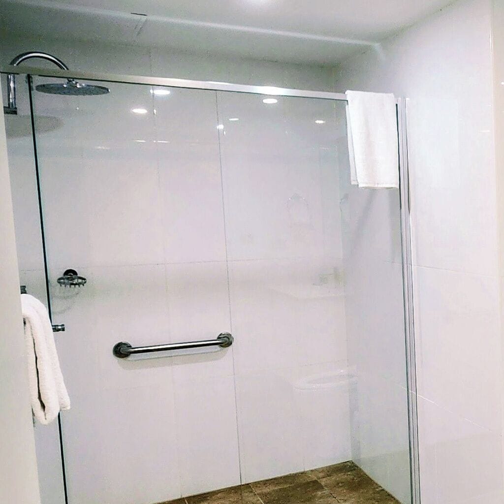 Una ducha con cerramiento de vidrio, barandilla, paredes de azulejos blancos y una toalla visible a la izquierda.